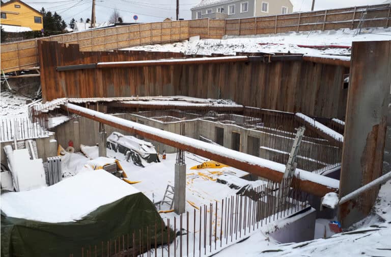 Støttemur under bygging i vintervær med snø, byggeutstyr og forskaling på byggeplass. Fokus på fundamentering og konstruksjon av sikre støttemurer.