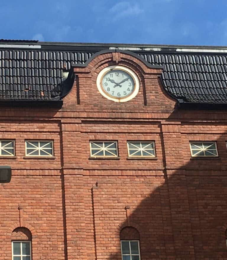 Gammel rødmalt bygning med klokketårn og utendørs klokke, arkitekturstil med murstein og vinduer, historisk byggverk i Norge for moderne nettside presentasjon.